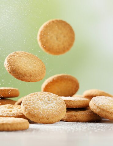 Golden round cookies piled and scattered on a white surface, fine sugar dust floating in the air.