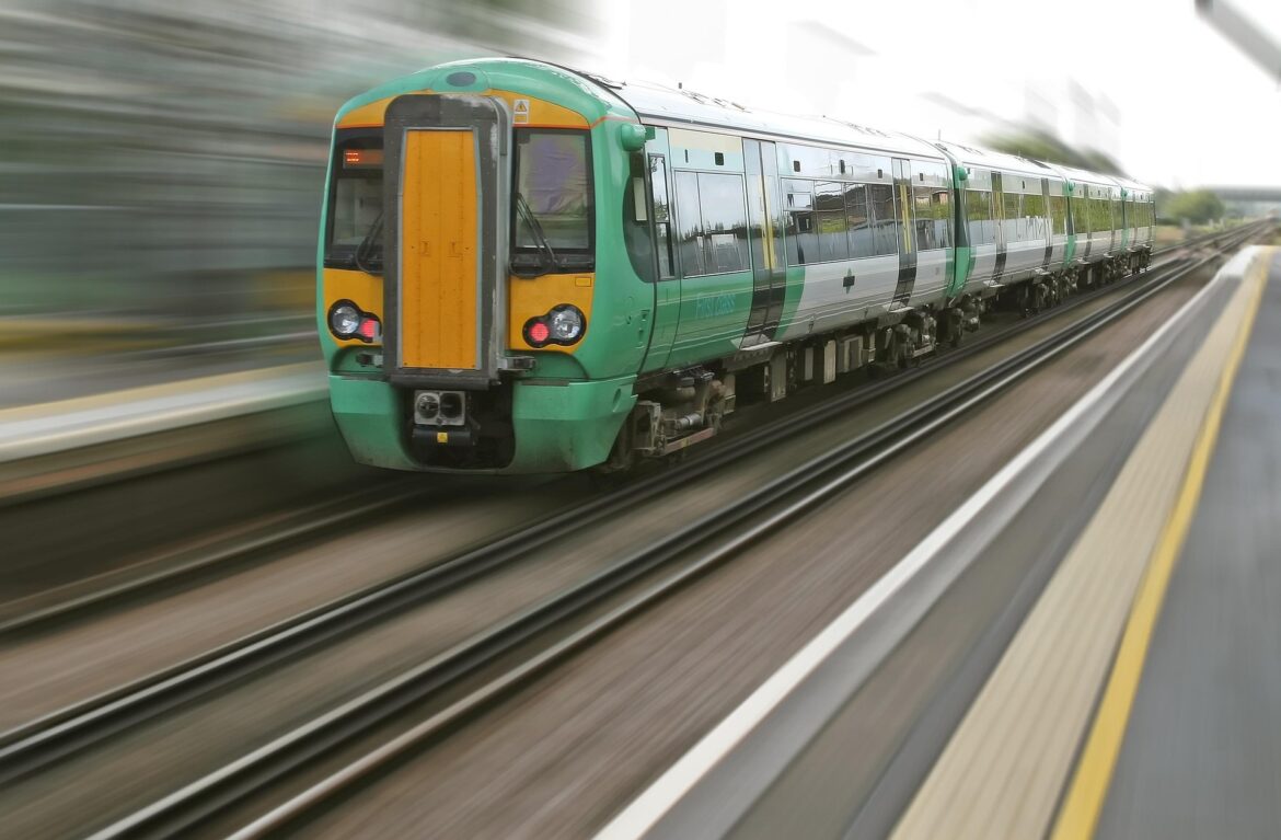 Green and yellow passenger train speeding along the tracks, foreground platform visible with motion blur in the background.