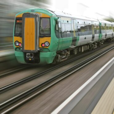 Green and yellow passenger train speeding along the tracks, foreground platform visible with motion blur in the background.