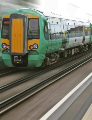 Green and yellow passenger train speeding along the tracks, foreground platform visible with motion blur in the background.