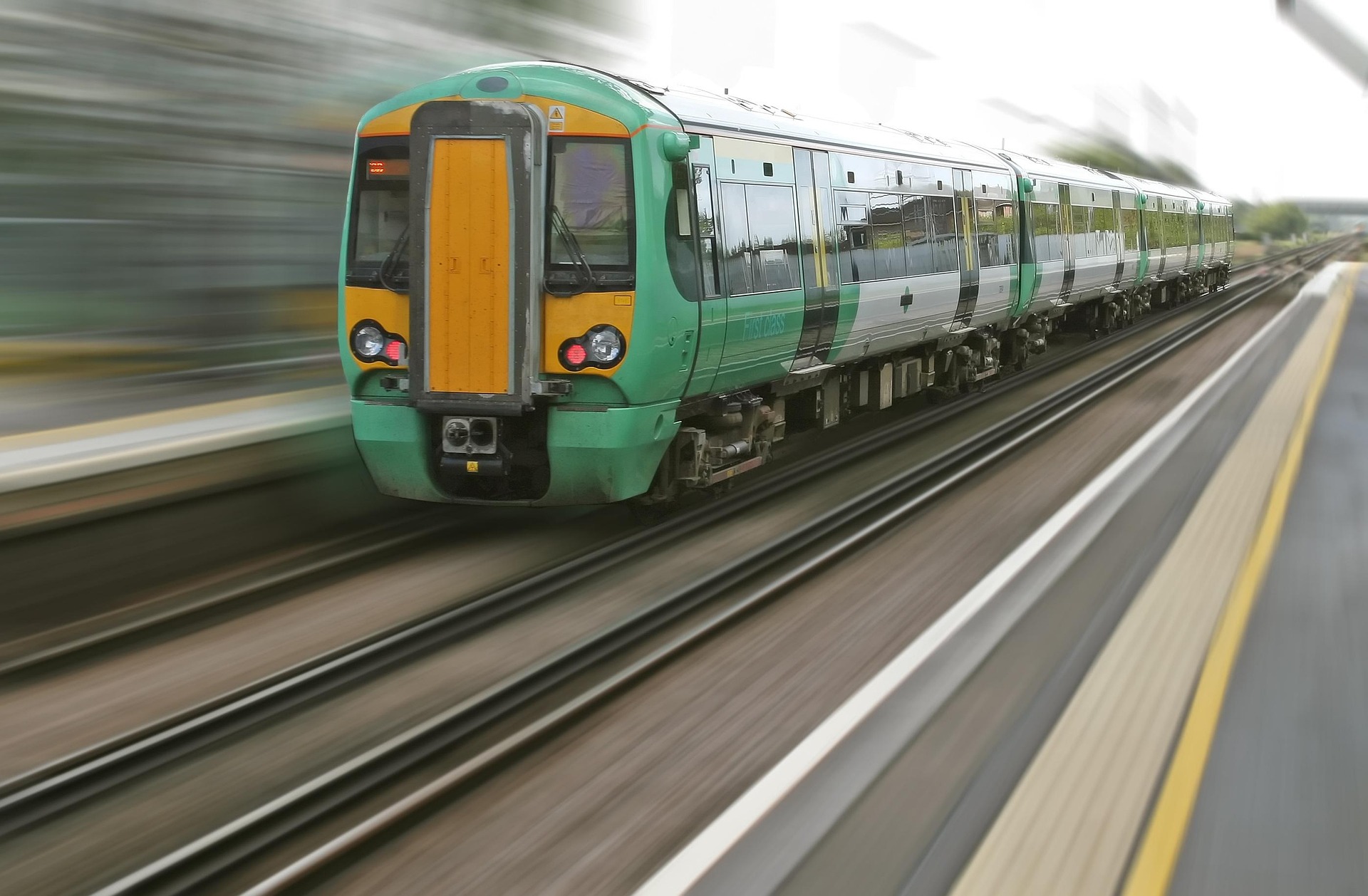 Green and yellow passenger train speeding along the tracks, foreground platform visible with motion blur in the background.