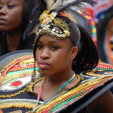 Young woman in a colorful carnival costume with gold beadwork, feathers, and a large decorative shield hold.