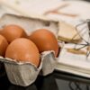 Brown eggs in a gray carton on a dark countertop, with a whisk and recipe pages in the background.