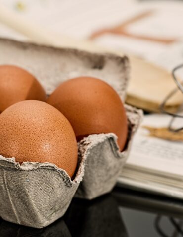Brown eggs in a gray carton on a dark countertop, with a whisk and recipe pages in the background.