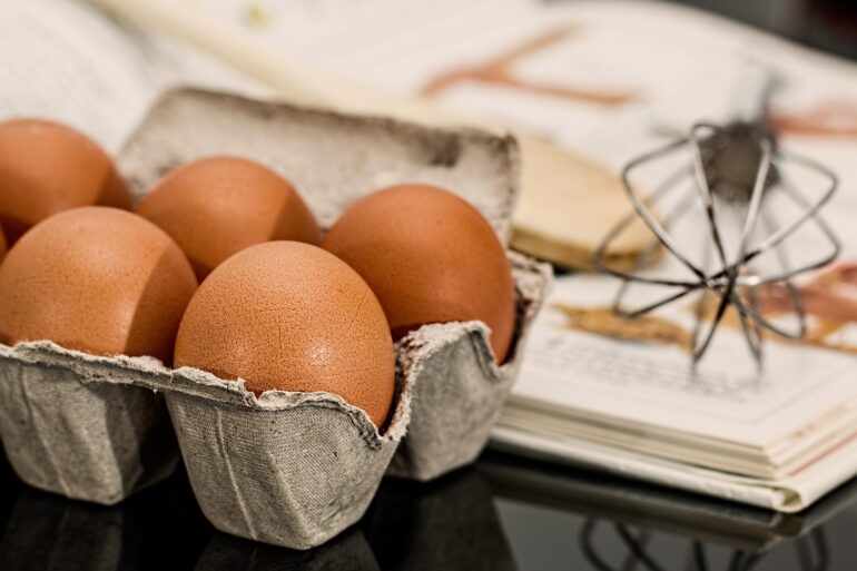 jajka - NOTE.radio - Polskie radio w UK | Polskie radio w Londynie Brown eggs in a gray carton on a dark countertop, with a whisk and recipe pages in the background.