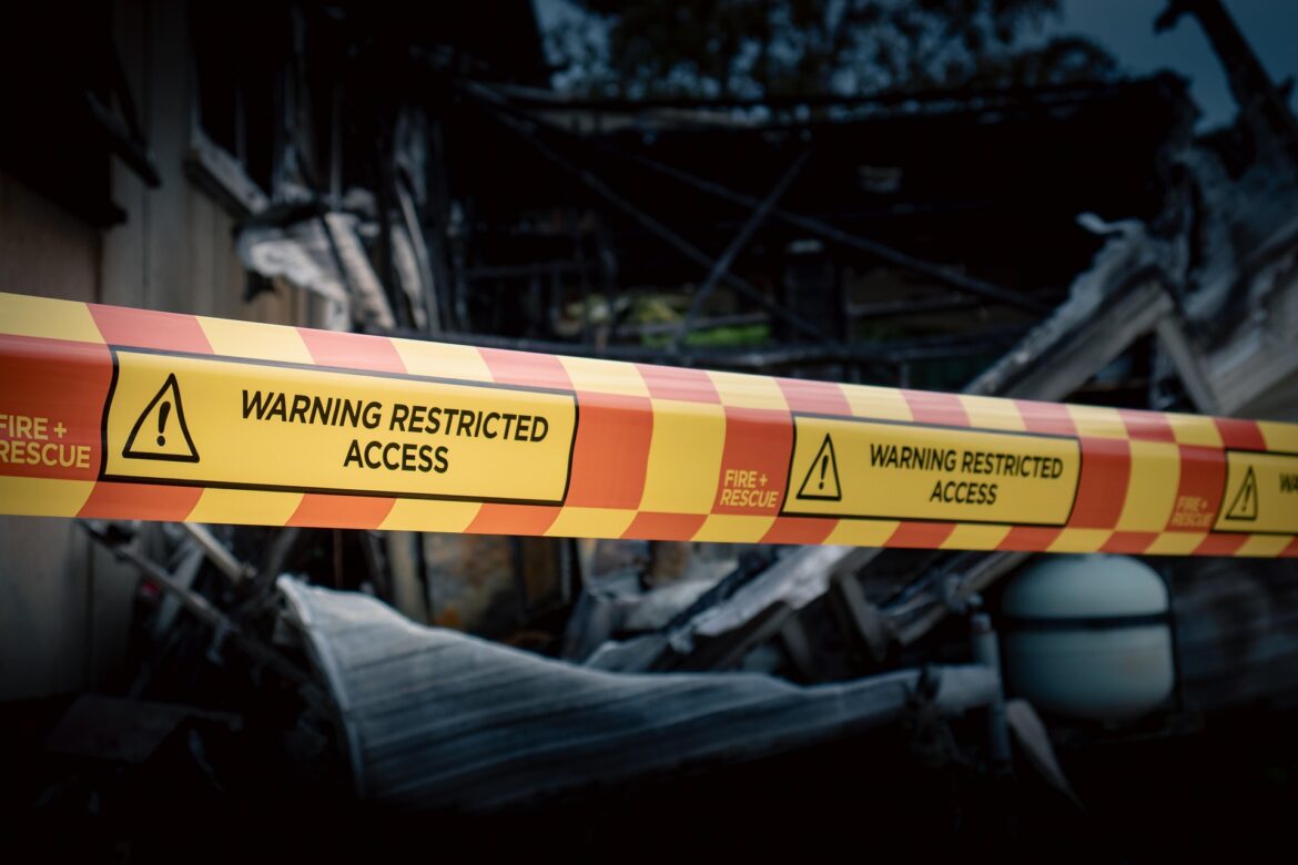 Yellow caution tape reading 'Warning Restricted Access' stretched across burned rubble, blocking a damaged building.