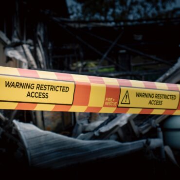 Yellow caution tape reading 'Warning Restricted Access' stretched across burned rubble, blocking a damaged building.