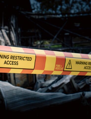 Yellow caution tape reading 'Warning Restricted Access' stretched across burned rubble, blocking a damaged building.