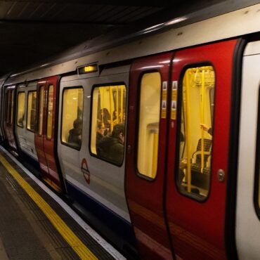 A London Underground train stopped at an underground platform, with passengers inside and a few people waiting on the platform edge.