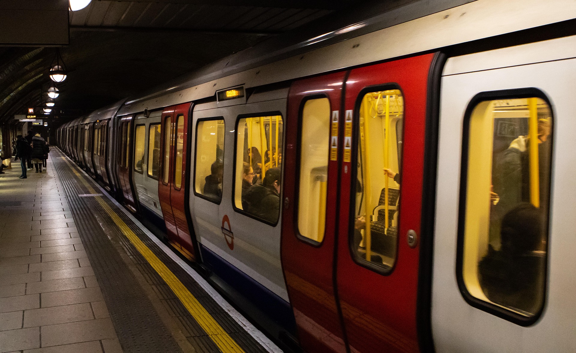 A London Underground train stopped at an underground platform, with passengers inside and a few people waiting on the platform edge.