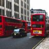 Red double-decker buses travel down a London street lined with tall gray buildings and a sidewalk.",