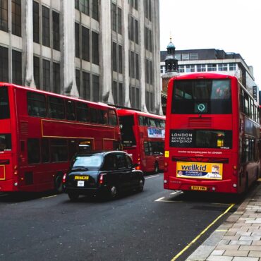Red double-decker buses travel down a London street lined with tall gray buildings and a sidewalk.",