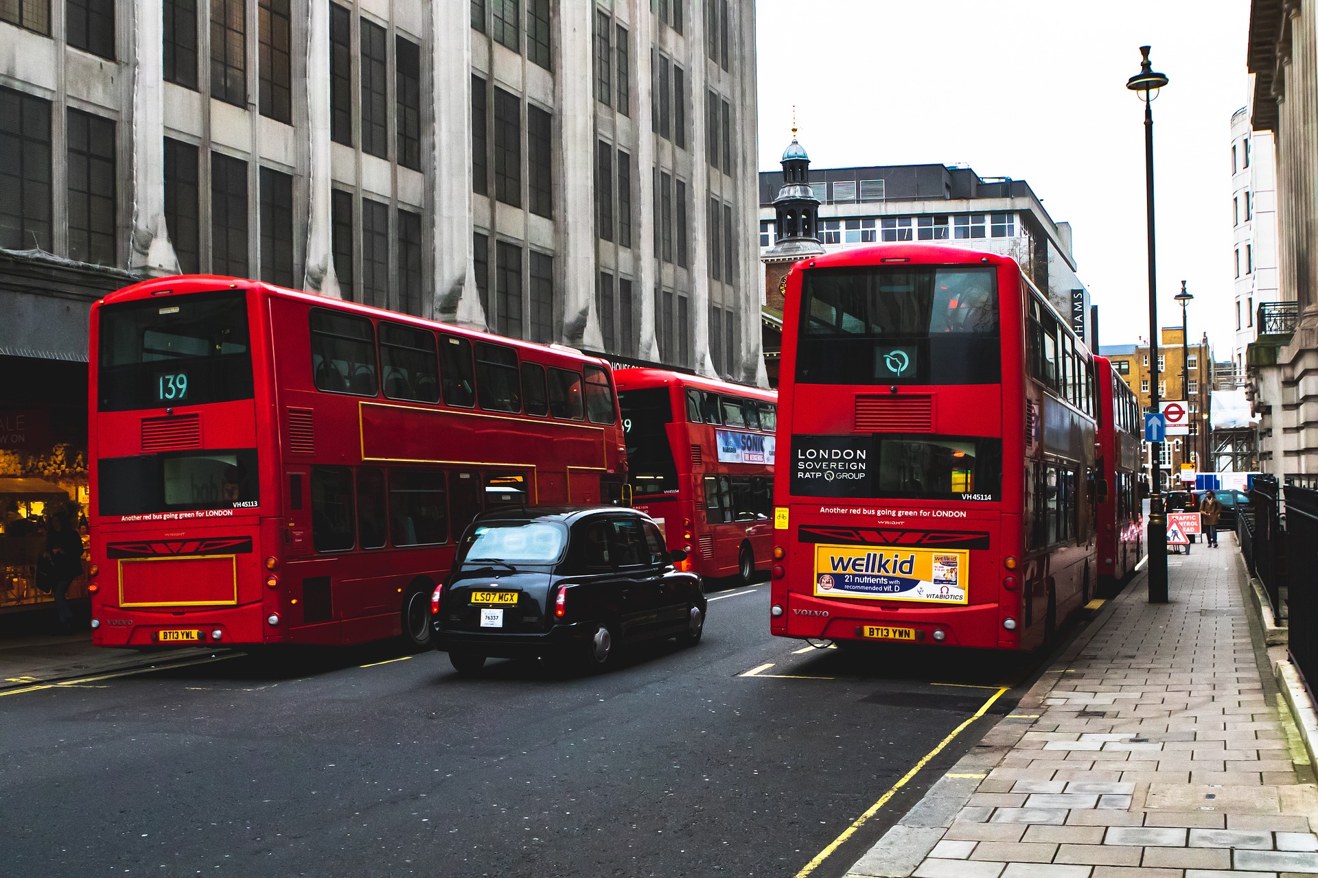 Red double-decker buses travel down a London street lined with tall gray buildings and a sidewalk.",