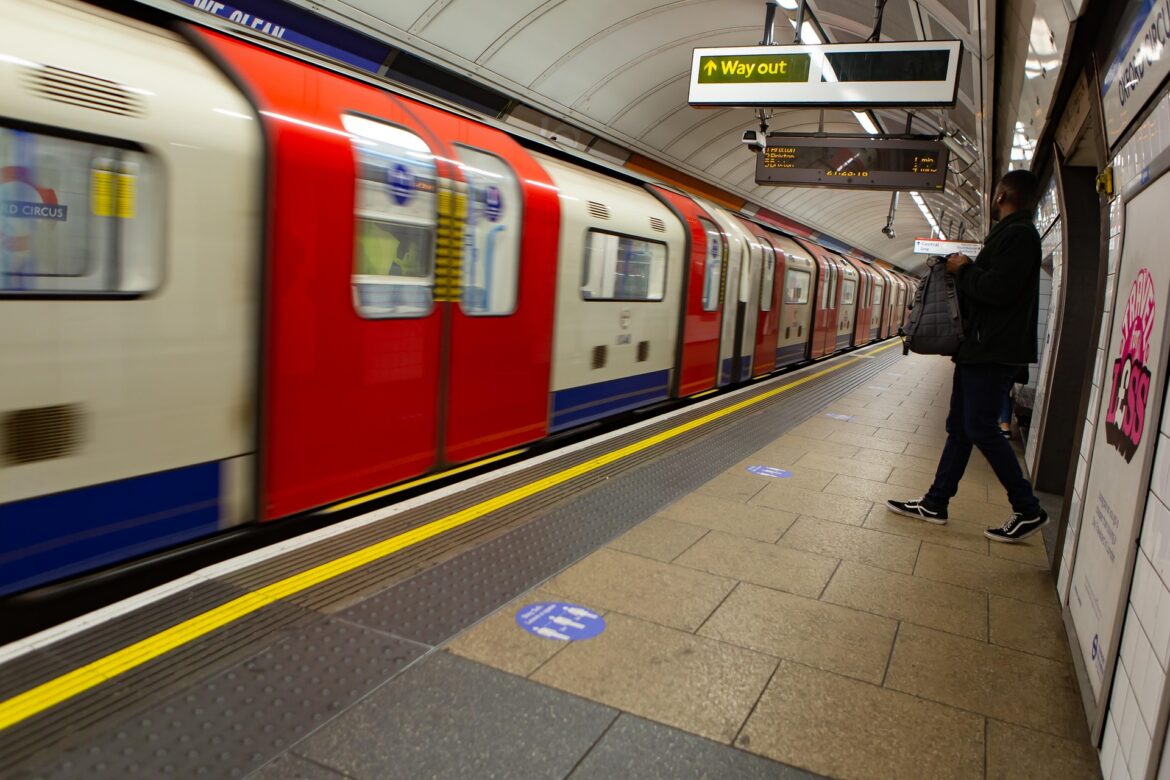 Man with backpack standing on a subway platform as a red and cream train passes by.