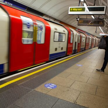 Man with backpack standing on a subway platform as a red and cream train passes by.