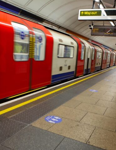 Man with backpack standing on a subway platform as a red and cream train passes by.