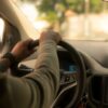 Person's hands grip the steering wheel from the driver's seat with a blurred dashboard and car radio in view, daytime light outside.