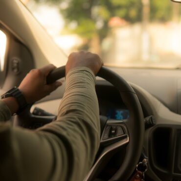 Person's hands grip the steering wheel from the driver's seat with a blurred dashboard and car radio in view, daytime light outside.