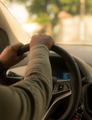 Person's hands grip the steering wheel from the driver's seat with a blurred dashboard and car radio in view, daytime light outside.