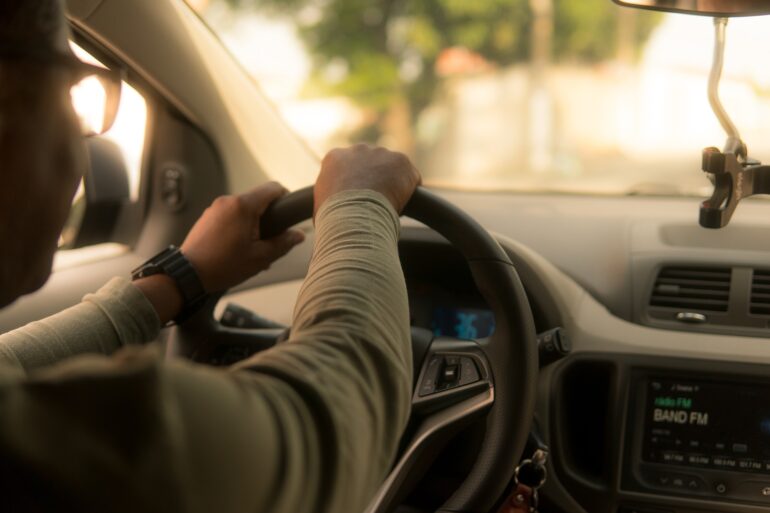 Person's hands grip the steering wheel from the driver's seat with a blurred dashboard and car radio in view, daytime light outside.