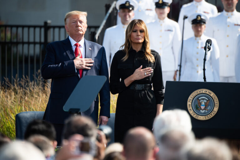 President Donald Trump and first lady Melania Trump stand with their right hands over their hearts during a ceremony, with uniformed military personnel behind them and the presidential seal on the lectern in the foreground.