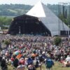 Huge outdoor music festival crowd gathered in front of a triangular-pyramid stage with large backup screens and scaffolding structures.