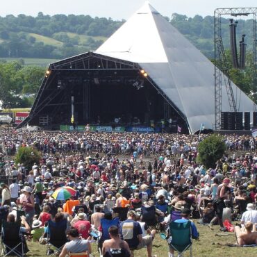Huge outdoor music festival crowd gathered in front of a triangular-pyramid stage with large backup screens and scaffolding structures.