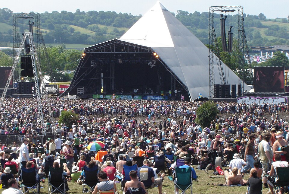 Huge outdoor music festival crowd gathered in front of a triangular-pyramid stage with large backup screens and scaffolding structures.