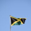 Jamaican flag waving on a flagpole against a clear blue sky, featuring a black X-shaped cross with gold edges and green triangles at the corners.