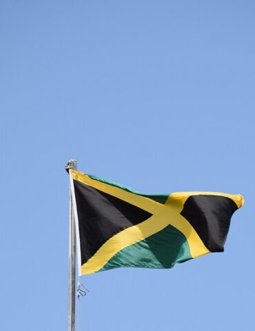 Jamaican flag waving on a flagpole against a clear blue sky, featuring a black X-shaped cross with gold edges and green triangles at the corners.