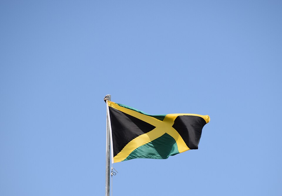 Jamaican flag waving on a flagpole against a clear blue sky, featuring a black X-shaped cross with gold edges and green triangles at the corners.