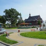 Circular paved plaza with benches and a roadside shop on the left, a small silver car parked nearby, a tall stone obelisk in the grass, and a wooden church in the background under a clear blue sky.