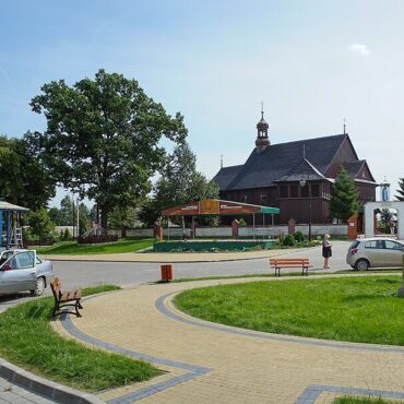 Circular paved plaza with benches and a roadside shop on the left, a small silver car parked nearby, a tall stone obelisk in the grass, and a wooden church in the background under a clear blue sky.