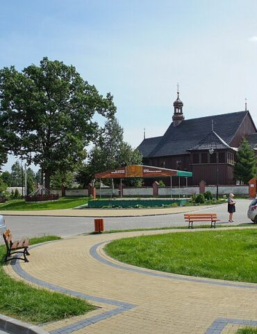 Circular paved plaza with benches and a roadside shop on the left, a small silver car parked nearby, a tall stone obelisk in the grass, and a wooden church in the background under a clear blue sky.