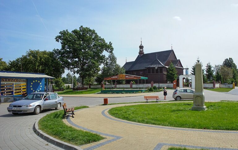 - NOTE.radio - Polskie radio w UK | Polskie radio w Londynie Circular paved plaza with benches and a roadside shop on the left, a small silver car parked nearby, a tall stone obelisk in the grass, and a wooden church in the background under a clear blue sky.