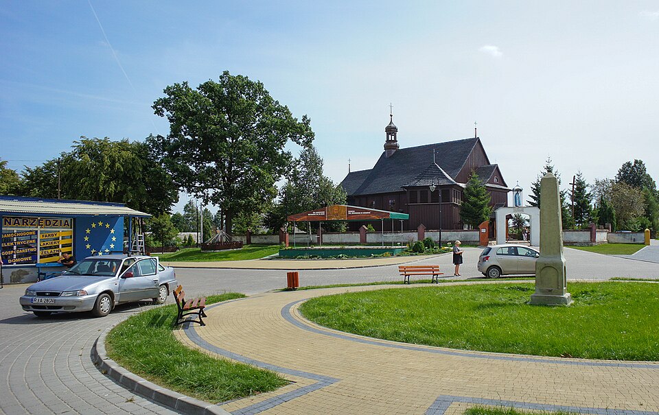 Circular paved plaza with benches and a roadside shop on the left, a small silver car parked nearby, a tall stone obelisk in the grass, and a wooden church in the background under a clear blue sky.