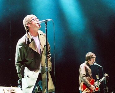 Lead singer in a beige coat performs at the mic on a dark stage; guitarist in the background under blue lights.3a4️ guitarist is playing in the background under blue stage lighting for a live concert.