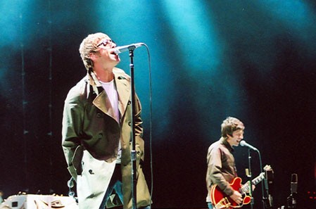 Lead singer in a beige coat performs at the mic on a dark stage; guitarist in the background under blue lights.3a4️ guitarist is playing in the background under blue stage lighting for a live concert.