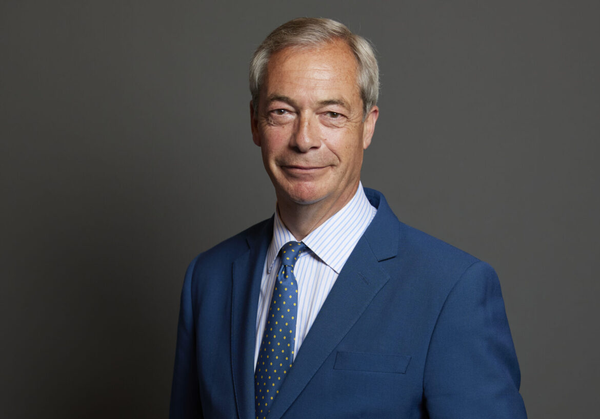 Smiling man in a blue suit and polka-dot yellow tie, head-and-shoulders portrait against a gray backdrop.
