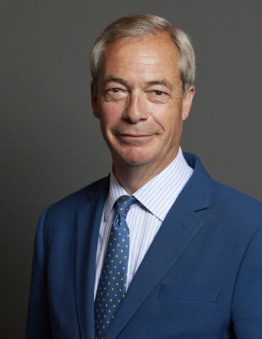 Smiling man in a blue suit and polka-dot yellow tie, head-and-shoulders portrait against a gray backdrop.