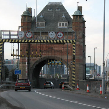 Brick gateway over a roadway leading to a tunnel, with height clearance signs above the lane and striped supports on either side.