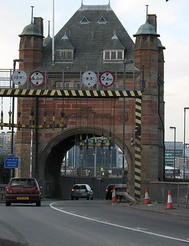 Brick gateway over a roadway leading to a tunnel, with height clearance signs above the lane and striped supports on either side.