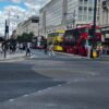 Busy city street with red double-decker buses, pedestrians, and modern glass-fronted buildings.