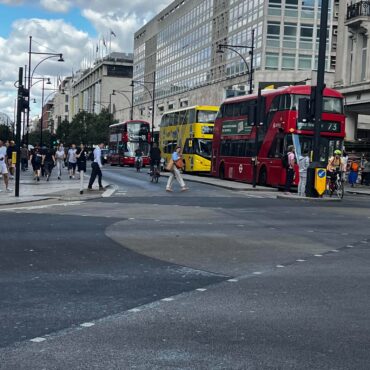 Busy city street with red double-decker buses, pedestrians, and modern glass-fronted buildings.