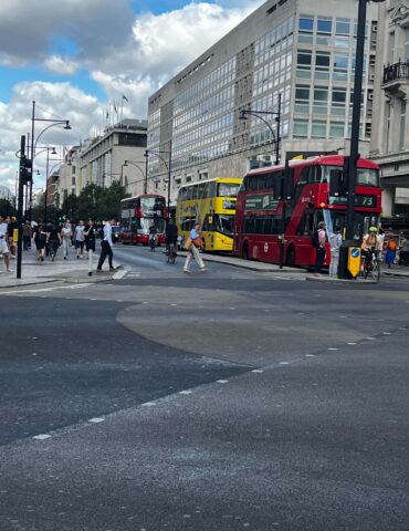 Busy city street with red double-decker buses, pedestrians, and modern glass-fronted buildings.