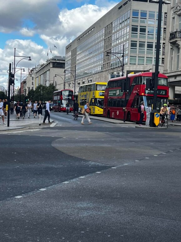 Busy city street with red double-decker buses, pedestrians, and modern glass-fronted buildings.