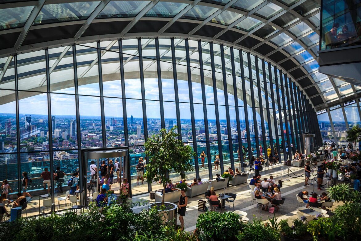 Glass-walled observation deck with city skyline, people seated at tables and walking among potted plants.