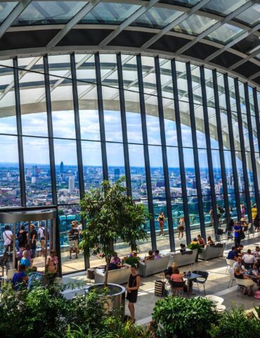Glass-walled observation deck with city skyline, people seated at tables and walking among potted plants.