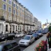 Row of classic Georgian townhouses with ornate window frames and black iron balconies, parked cars lining a sunny street. Flowers along the sidewalk railings.