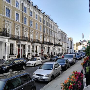 Row of classic Georgian townhouses with ornate window frames and black iron balconies, parked cars lining a sunny street. Flowers along the sidewalk railings.